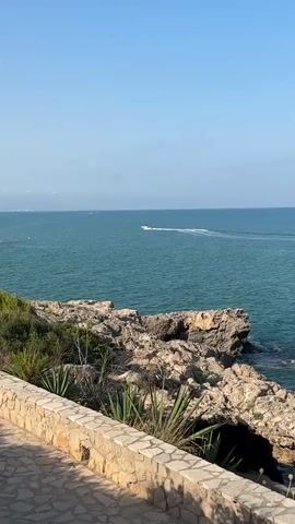 A vertical video shows a view of the sea from a rocky coastline. The water is a vibrant turquoise color, and the sky is clear blue. In the distance, a white speedboat is visible, leaving a wake as it moves across the water. The foreground features a stone wall and a paved walkway, with sparse vegetation growing on the rocky outcrop. The sun is shining, casting a warm light on the scene. The overall atmosphere is peaceful and serene.