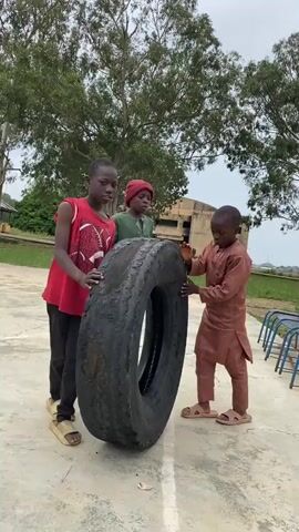 Three young boys are gathered around a large, worn tire on a paved surface. The boy on the left, wearing a red sleeveless shirt and black pants, is positioned facing the camera with his hands on the tire. The middle boy, in a green outfit and red head covering, also has his hands on the tire. The boy on the right, dressed in a brown robe, is on the other side of the tire, pushing it with his hands. The background shows trees, a building, and a basketball hoop. The lighting suggests an outdoor environment during the day.