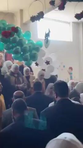 The scene depicts a crowded indoor gathering, potentially a graduation or award ceremony. A group of people, predominantly women wearing hijabs, are clustered together. One woman in the foreground is holding a rolled-up document, presumably a diploma or certificate. The crowd is a mix of men in suits and other individuals. Balloons in green and white, adorned with red star accents, decorate the space, adding to the celebratory atmosphere.  An emblem featuring an eagle design is visible in the background, potentially indicating the institution or organization hosting the event. The general setting suggests an educational or communal event within an institution.