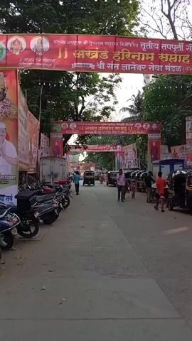The video shows a street in India with decorations hung across it for a religious festival.  Several banners in red and orange with text in Hindi frame the street. On the left side of the road, several motorcycles are parked, while on the right side, several auto-rickshaws, or tuk-tuks, are parked.  People are walking along the street, including one young person with a backpack walking toward the camera. An auto-rickshaw passes along the road, and a parked delivery truck is also visible. The street is paved and seems to be a local road in an urban or semi-urban area. The weather appears to be sunny. Trees are visible along the sides of the street.