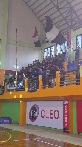 A crowd of spectators is visible in a gymnasium, standing and seated on bleachers. Several individuals are holding flags, with one large black and white flag prominently displayed. The spectators are predominantly wearing dark clothing. Below the bleachers, a large banner with the "Cleo" logo and text is visible, along with advertisements for "Biznet" and "IFL Indonesia". The gymnasium has a wooden floor, and the walls are painted green and yellow. Natural light is entering through windows at the top of the structure.