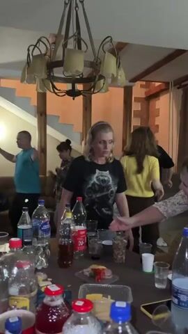 This media file captures a lively gathering indoors, possibly a party or a casual get-together. The scene is set in a room with rustic wooden accents, including the ceiling beams and support columns, and a prominent, ornate chandelier hanging overhead. A staircase with wooden balusters is visible in the background.

Several people are present, engaged in various activities. In the foreground, a table is laden with bottles of drinks (water, soda, and possibly other beverages), plastic cups, and some food items, including what appears to be a charcuterie or snack tray with cheese, meat, and vegetables.

The main focus in the mid-ground is on a group of individuals dancing and interacting. A woman in a black t-shirt with a graphic of a raccoon is looking towards the camera with a smile. To her left, a woman in a plaid shirt and another in a green top are also dancing. To the far left, a man in a teal t-shirt stands and looks away. Further back, another woman in a yellow top is visible. The overall mood seems joyful and energetic, suggesting a celebration or a social event.

The lighting indicates it is likely evening or indoors, with artificial light sources being dominant. There are no clear indications of the weather or time of day outside. The mention of "Zavod Mikhailovskii, Russia" as a location context provides geographical information, placing the event in a specific region of Russia.

There are no visible brand names or text on the clothing of the individuals, but some labels are visible on the drink bottles. One bottle has "Аква Минерале" (Aqua Minerale) clearly legible. Another bottle has what appears to be Russian text. The t-shirt worn by the woman in the foreground has a graphic of a raccoon.
