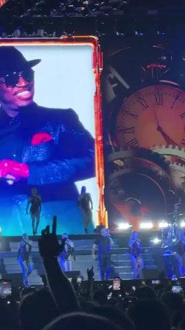 A live performance takes place on a stage with a large screen displaying a close-up of a man's face wearing a hat and sunglasses. Below the screen, several dancers in revealing outfits perform on elevated platforms. In the center of the stage, a bald man in a black suit sings into a microphone, accompanied by the dancers. To the right of the screen, a large, vertical display shows a tower of rotating blocks with letters on them, some of which light up green to form the word "LIVES." The background of the stage features a large clock face with visible gears, suggesting a steampunk theme. The audience is visible in the foreground, with many people holding up their phones to record the performance. The stage is brightly lit with blue and orange lights, and confetti appears to be falling from above.