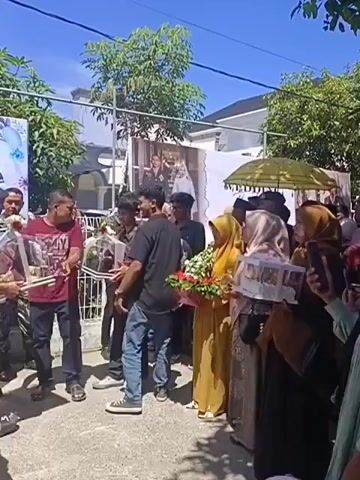This video shows an outdoor gathering of people, possibly a wedding procession or celebration, under a bright sky. Several individuals are holding decorative items, including bouquets of flowers and what appear to be birdcages containing small birds. A large banner or backdrop featuring a couple in formal attire is visible behind the crowd. Some people are dressed in traditional religious clothing, such as hijabs. The scene suggests a festive occasion with an exchange of gifts or symbolic items.