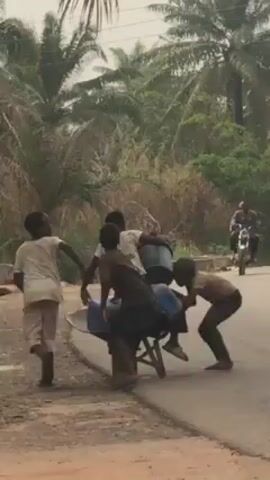 The video depicts a group of children pushing a wheelbarrow filled with blue plastic containers along a paved road in a rural area with lush vegetation. One child carries a container on his head, while others push and pull the wheelbarrow. A person rides a motorcycle on the road. The children appear to be working together, and their expressions are difficult to discern. The environment consists of palm trees and other dense greenery.