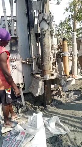 The video shows two men working on a well-drilling machine in an outdoor environment. One man, wearing a purple head covering, an orange sleeveless shirt, and black shorts, stands to the left of the machine, seemingly operating it or monitoring its progress. He holds a thin metal rod in his hand. The second man, wearing an orange patterned shirt and dark shorts, is on the right side of the machine, manipulating a wooden object. The machine itself is a large, heavy-duty drilling rig, predominantly gray in color, with various pipes, levers, and gauges visible. The ground around the machine is a mixture of dirt and gray sludge. Several opened bags, possibly containing drilling materials, are scattered on the ground near the base of the machine. In the background, there are trees and vegetation, suggesting the scene is in a rural or semi-rural area.
