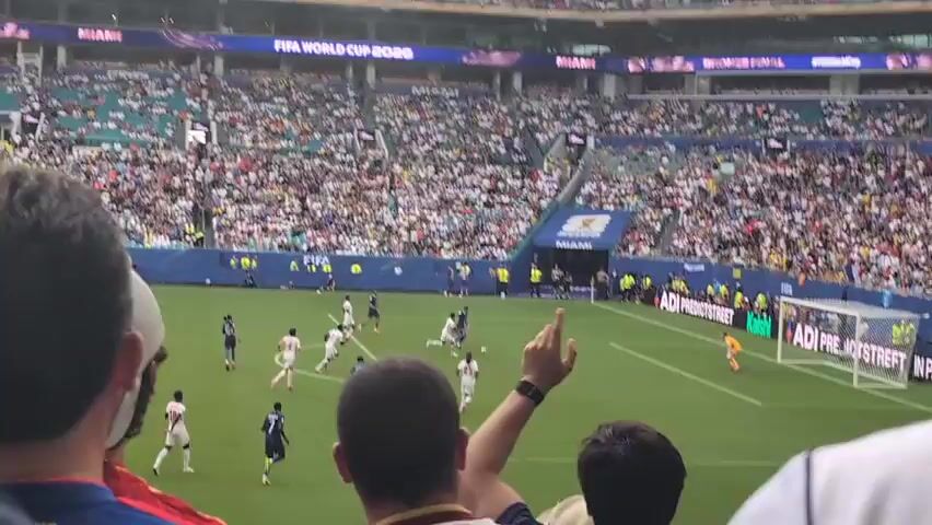 A soccer game is being played in a crowded stadium. Several players are on the field, some wearing white and others wearing blue. One player in blue is running towards the goal, while another player in white is defending. A goalkeeper in orange is in position in front of the net. Spectators fill the stands, many of them wearing white shirts. The stadium has signs that read "FIFA WORLD CUP 2026" and "MIAMI". The field is green with white lines marking the boundaries. The action appears to be near the goal, and the crowd seems engaged.