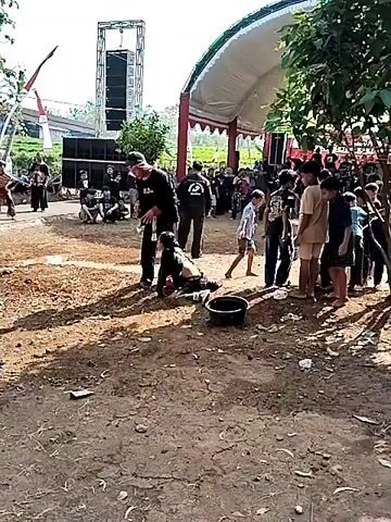 In an outdoor setting with a dirt ground and some vegetation, a group of people are gathered. In the foreground, a man in a black shirt and pants is kneeling on the ground, possibly in a performance or demonstration. Another person, also in dark clothing, is standing over him, appearing to guide or assist. A black bucket is placed near the kneeling person. In the background, there are speakers and sound equipment, suggesting an event like a concert or festival. Several other individuals are standing around, observing the scene. The overall atmosphere appears to be a public gathering with some form of entertainment or ritual taking place.
