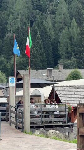The video shows a group of people sitting at a table on an outdoor deck. They are seated on benches and appear to be eating or drinking. Two flags are visible, one blue and yellow and the other the Italian flag. The background consists of a dense forest of pine trees, and there are buildings with sloped roofs visible. There is a wooden railing in the foreground, and a paved walkway curves away from the camera.