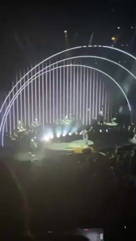 The video shows a concert with a large crowd of people, viewed from the perspective of someone within the audience. A single performer stands on a raised platform on the stage, surrounded by an elaborate lighting setup including multiple colored spotlights, vertical lights, and arched neon structures. Smoke or fog effects are also visible. The audience is actively engaged, with many individuals raising their hands and recording the performance on their phones. The lighting creates a dynamic and colorful atmosphere, shifting between various hues of purple, blue, green, yellow, and orange.