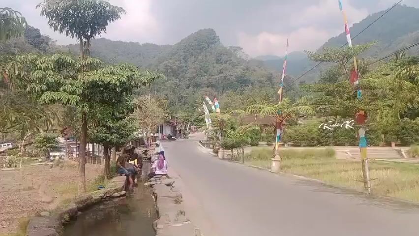 A wide shot shows a rural road in a valley with lush green mountains in the background. Several people are gathered near a narrow water channel on the left side of the road. Two men are sitting by the channel, with one of them dipping his feet in the water. Another person, possibly a woman, is standing next to them. The road is paved and appears to be a main thoroughfare, with flags on poles lining the side. Buildings and trees are visible along the road, suggesting a small village or community. The overall scene depicts a peaceful, everyday moment in a natural setting.