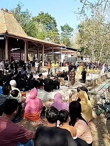 The video shows an outdoor gathering in front of a building with a traditional tiled roof, possibly a temple or shrine. Many people are present, with a group sitting on the ground in the foreground, facing a raised area where a performance or ceremony is taking place. Several individuals, some dressed in traditional attire including patterned fabrics and headwear, are engaged in an activity on the raised platform. Others are standing or sitting around this central area. The environment appears to be somewhat rural, with trees and natural ground visible. The lighting suggests it is daytime, likely with bright sunlight. The overall atmosphere seems to be one of cultural observance or community event.