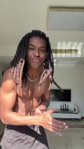 A young man with dreadlocks, wearing only black shorts, bounces a soccer ball on his leg in a living room. The room is modern, with white walls, a beige and brown tiled floor, a white entertainment console, a flat-screen TV, and a white ceiling light. There is a closed doorway to the left of the frame, and a clock on the wall. Music is playing in the background.