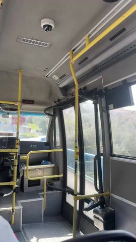 The camera is situated inside a bus, pointed forward, capturing the interior from near the driver's area towards the front door. Yellow handrails are prominent, lining the aisle. To the left is the driver's area with a blue patterned seat visible. Outside the front windows, one can see a bridge with blue railings and green foliage in the distance. A security camera is mounted on the ceiling of the bus. The vehicle appears to be moving.