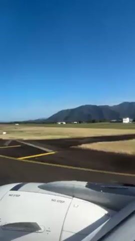 The view from an airplane window shows an airport runway on a sunny day. The wing of the plane is visible in the foreground with "NO STEP" printed on it. The runway is black with yellow markings. Beyond the runway is a grassy field, and in the distance, there are mountains. The sky is clear and blue. There are a few buildings visible in the distance as well. The plane appears to be stationary on the runway.