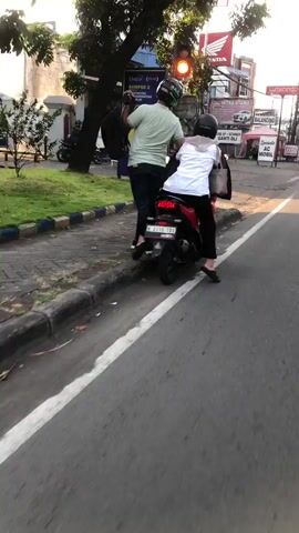 Two people are riding a motorcycle on a street. The person in front, wearing a light green t-shirt and dark pants, is driving the motorcycle. They are wearing a black helmet. The person in the back is wearing a white shirt and dark pants, also wearing a black helmet. They are carrying a black bag behind them. The motorcycle is red and black.

The scene appears to be in Gelam, Indonesia, as indicated by the location context. The time of day is likely late afternoon or early evening, given the warm, golden light and long shadows. The weather appears to be clear. The motorcycle is on a paved road with a white line marking a lane. To the left of the road, there is a sidewalk with grass and a curb. In the background, there are buildings, trees, and street signs. One sign reads "MITRA UTAMA" and another says "AC MOBIL." A traffic light is visible in the distance, showing a yellow light. The overall mood is neutral, capturing a moment of everyday transportation.