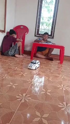 Two young boys are in a room. One boy is sitting on the floor and playing with a toy car. The other boy is sitting at a red table and appears to be drawing or writing. There is a red chair in the background. The room has tiled flooring and windows with light coming through them.