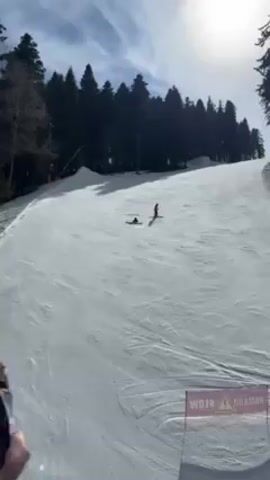The video shows a snowy ski slope on a sunny day with trees in the background. A skier wearing dark clothing and light blue pants is sitting on the snow. Another skier is standing further up the slope. A sign that says "SLOW" with an exclamation point is visible at the bottom of the frame.