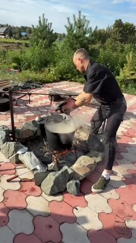 A man wearing a black shirt, black pants, and green sneakers with oven mitts on his hands is in the process of moving a large black pot from a fire pit. The fire pit is made of rocks and contains a fire with flames. Two metal ladles are placed on the rocks around the fire. The pot is steaming, indicating it contains hot liquid. The man is lifting the pot and appears to be walking away from the fire pit. The background shows a rural outdoor setting with green bushes, trees, and a blue sky with some clouds. The ground around the fire pit is paved with interlocking red and white tiles. There is also a metal grill or stand to the left of the fire pit.