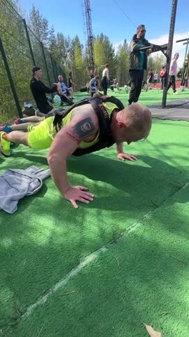 A medium shot captures three men doing push-ups on an outdoor turf area during a competition. The main subject is a muscular man in the foreground with blond hair, wearing a black weight vest over a neon green tank top and neon green shorts with matching shoes. He has a tattoo on his upper left arm. To his left is a gray sweatshirt. Another man in a purple shirt and black shorts performs push-ups next to him. In the background, a man stands near a metal structure, possibly a pull-up bar, taking notes. A chain link fence surrounds the area, with trees visible behind it. A tall metal tower and other competitors and spectators are visible further in the background. The sky is blue with scattered clouds. The overall scene shows an outdoor fitness competition.