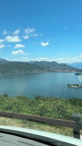 This is a video of a scenic lake with islands, taken from a moving vehicle. The water is a deep blue, and the sky is clear with a few white clouds. Lush green trees and foliage are in the foreground, and mountains can be seen in the distance. There are a few boats on the water, and buildings are visible on the shore and on the islands. The camera pans slowly to the right, offering a wider view of the landscape.