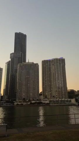 The video shows a panoramic view of a city skyline at dusk, with several tall buildings reflected in a body of water. The sky is a soft gradient of orange and grey, indicating the sun is setting. In the foreground, there is a grassy area with a park bench and a few people sitting. The water appears calm, with a small boat visible in the distance. The overall atmosphere is peaceful and serene.
