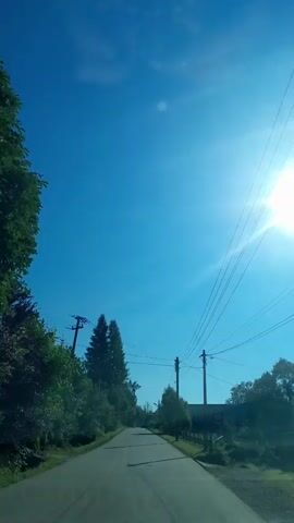 The video shows a first-person perspective of driving down a paved road on a sunny day. The sky is a clear, bright blue with the sun prominently visible and causing lens flare. On either side of the road, there are trees and foliage. Utility poles with wires are visible along the roadside. The road appears to be in a rural or semi-rural setting.