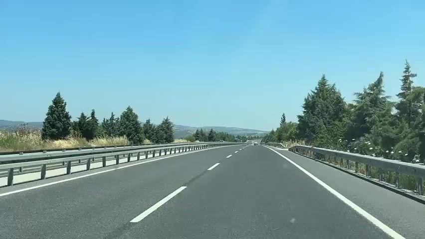 The video shows a first-person perspective driving on a highway. The road is paved, with clear white lane markings. On either side of the highway are guardrails, followed by grassy areas and numerous evergreen trees. The sky is clear and blue. There are distant hills and some smaller vehicles visible on the road ahead. The sun appears to be shining brightly, suggesting it is daytime. The camera perspective is fixed, indicating it is likely mounted on the dashboard or windshield of a vehicle. There are no people visible within the immediate vicinity of the vehicle. The environment appears to be rural or semi-rural.