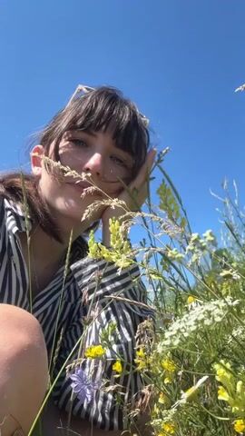 A young woman with dark hair and bangs, wearing sunglasses perched on her head and a striped shirt, crouches in a field of wildflowers under a clear blue sky. She is interacting with the flowers, bringing some to her face and then covering her face with her hand, smiling broadly and revealing braces. The focus is on her face and the surrounding plants, with the background being the sky.
