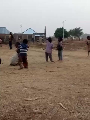 Several children play in a dry, grassy area near simple buildings. Two children are in the foreground wrestling on the ground near a tire. Two other children are standing facing each other. Other individuals stand and walk in the background. There is a utility pole and a small tree visible as well. The sky is overcast and gray.