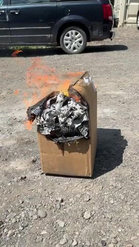 A cardboard box is burning in a gravel lot. The box is mostly intact and upright, with flames and smoke coming from the top. The contents of the box are charred and appear to be paper or similar material. In the background, a dark-colored van is parked. The ground is covered in gravel and some debris. The lighting suggests it is daytime.
