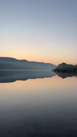 The video shows a calm lake at dawn. The sky is a gradient of pale blue at the top transitioning to a soft orange and pink near the horizon. Mist hovers over a distant, silhouetted mountain range. The water is perfectly still, reflecting the sky and the land with mirror-like clarity. A small, tree-covered island is visible in the middle ground, also reflected in the water. The foreground shows the dark, rocky bottom of the lake. The overall atmosphere is serene and peaceful. There are no people or animals visible in the video, and no discernible actions or interactions are taking place.