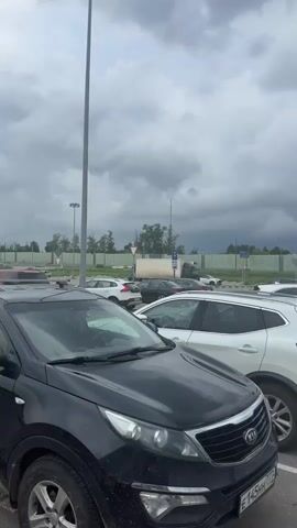 The video shows an outdoor parking lot with several cars. The sky is overcast with dark clouds, suggesting an impending storm. A parking sign is visible in the foreground. In the middle ground, two men are interacting with a silver car. One man, wearing a white shirt and a cap with a "Z" logo, is standing on the roof of the car. The other man, wearing a green t-shirt, is standing next to the open driver's side door of the car. The scene appears to be a casual setting, possibly a parking lot for a shopping center or event.