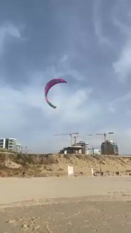 The video shows a colorful kite flying over a beach on a cloudy day. The kite is teal, pink, and yellow and is shaped like a crescent moon. In the background, there are buildings and construction cranes. The beach is sandy and there are a few people walking along the shore. There is also a sign planted in the sand. The camera angle is from below, looking up at the kite. The kite moves around in the air.