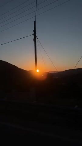 The video captures a serene sunset over a landscape featuring hills and a body of water. The sun, a bright orange orb, is prominently positioned near the horizon, casting a warm glow and creating lens flares on the camera. The sky transitions from a deep twilight blue at the top to vibrant orange and yellow hues around the setting sun.

The foreground is dark and largely in silhouette, featuring the top of a guardrail and a power pole on the left side. Beyond these, dark hills or mountains frame the central view of the sunset and the water body, which is also reflecting the sun's light. The overall scene is peaceful, showing the natural beauty of the sunset from what appears to be an elevated vantage point. No people or visible text are present in the video. The setting aligns with a coastal or mountainous area like Kaş, Turkey.