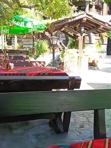 The video shows an outdoor seating area of a restaurant or cafe. In the foreground, there is a wooden table with a red, green, and yellow striped tablecloth. A dark green wooden bench is visible in front of the table. In the mid-ground, there are more tables and benches, some covered by green umbrellas with a Heineken logo. There is a wooden structure with a roof that appears to be a gazebo or part of the building. Two wooden barrels filled with white flowers are placed near the tables. In the background, there is a multi-story building with wooden balconies and stone walls. The ground is paved with grey stones. The lighting suggests it is daytime, with bright sunlight casting shadows. There are no people visible in the video.