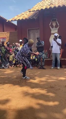 A group of people are gathered in a dusty, outdoor area. Two individuals, one wearing a purple and black patterned garment and the other in a blue and white patterned outfit, are dancing in the center. A crowd of onlookers, dressed in various colorful traditional African clothing, observe the dance. Some people are seated on the ground, while others stand. In the background, a building with a red facade and a tiled roof is visible, featuring a mural of a woman carrying a pot. A man in a white shirt and a white cap is standing to the right, looking at his phone, while another person is taking photos. The ground is covered in orange-red dirt. The sky is blue and clear, suggesting it is daytime.