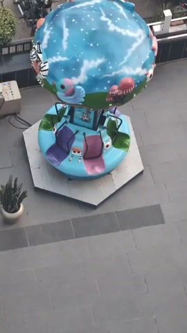 The video shows a children's amusement ride designed to look like a spinning globe. The globe is decorated with a starry sky and various cartoon characters. Below the globe is a circular platform with four seats, each colored differently (purple, green, pink, and blue). These seats are attached to the central structure and appear to swing outwards as the ride spins. The ride is set up on a paved area, possibly an outdoor plaza or sidewalk, with a control panel visible to the left and some parked motorcycles and scooters in the background. The ride is stationary at the beginning of the clip and then begins to slowly rotate.