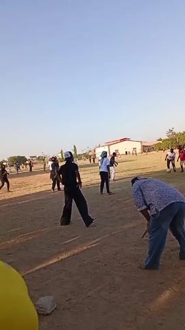 The video shows a group of people on a dirt field under a clear, bright sky. In the background, there is a building and some sparse vegetation. In the foreground, a person in a yellow shirt faces the camera briefly before turning to watch the activity on the field. There are numerous individuals scattered across the field, some walking and others appearing to be participating in an activity. They are dressed in a variety of casual clothing. The camera holder is near the ground, and a small rock is visible in the lower part of the frame. There are no clear interactions between the people.