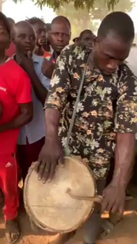 The video shows a group of people, mostly men, gathered outside in a somewhat sandy or dusty environment under some trees. The main focus is on a man in the foreground playing a small, light-colored drum, held at waist level by a strap around his neck. He is wearing a dark shirt with a floral pattern. The people around him are watching and listening. Some are looking directly at the drummer, while others are looking around. One man is wearing a red tracksuit with a "CHOICE" logo on the shirt, while another is wearing a blue shirt. A few people are wearing head coverings. The lighting appears to be natural daylight.