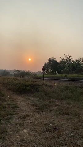 The video shows a sunset over a rural landscape. A dirt path is visible in the foreground, with dry grass and some small plants. In the middle ground, there are railroad tracks and a signal post. Beyond the tracks, there is a line of trees and vegetation. The sun is a bright orange disc in the hazy sky, casting a warm glow over the scene. The overall impression is one of tranquility and natural beauty.