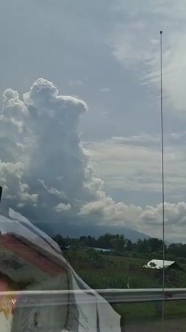 This video captures a scenic view from a moving vehicle. The sky is dominated by large, puffy white clouds, with one particularly tall and imposing cloud formation in the center left. In the distance, a range of mountains can be seen, partly obscured by the clouds. Below the sky, a lush green landscape unfolds, featuring fields and scattered trees. A white roof of a building is visible amidst the greenery. A highway barrier runs along the bottom of the frame, and a thin, vertical antenna extends upwards on the right side of the image. In the foreground, the corner of some papers or a book is visible, resting on a surface, possibly a car dashboard or window. A yellow and black road sign with a curved arrow indicating a sharp turn to the left is partially visible in the lower left quadrant. The overall impression is of a journey through a rural, possibly tropical, setting under a dynamic sky.