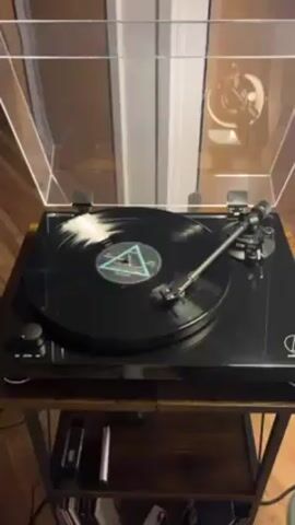 The video shows a black Audio-Technica turntable in operation. A vinyl record with a turquoise triangle design in the center is playing. The turntable is set up on a small dark brown and black metal shelving unit. The dust cover of the turntable is open. There are a few small electronic devices on the shelf of the unit, and several vinyl records are stacked on the bottom shelf. The floor is wood, and a white paneled wall is visible in the background. A partially visible light fixture is reflected on the clear dust cover of the turntable.