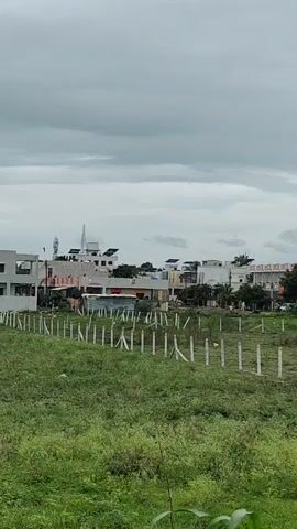 The video shows an outdoor scene with a partially built structure or empty lot in the foreground. A fence made of concrete posts and wire runs along the edge of the lot. In the background, there are several houses, some of which appear to be under construction or recently built, with white walls and windows. Some houses have solar panels on their roofs. Trees and greenery are scattered throughout the scene. The sky is overcast and gray, suggesting cloudy weather. There are power lines and poles visible in the video. The perspective changes slightly throughout the short clip, moving from a wider view of the landscape to a closer look at trees and then back to the houses. No people are visible in the video.