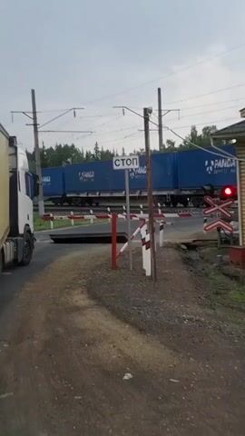 A train carrying blue shipping containers is crossing a railroad track that intersects a dirt road. A white truck is stopped on the left side of the road, waiting for the train to pass. A railroad crossing gate is down, and a stop sign is visible. The sky is overcast, and there are trees in the background. The time stamp at the beginning of the video is 00:00.
