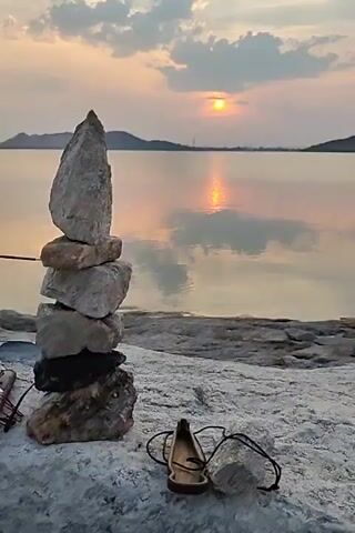 The camera remains still, focusing on a serene landscape. In the foreground, a pile of gray and black stones stacked high stands to the left of the frame. Near the base of the stones lies a wooden sandal and a coil of wire. The ground beneath these objects is white and textured.

Behind these objects, the eye is drawn to a calm body of water reflecting the golden hues of the setting sun. The sun, a small, bright orb, is positioned above distant mountains, adding depth to the scene. The sky is partly cloudy, with the colors of the sunset subtly blending across the horizon.