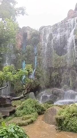 A man is walking along a path towards a waterfall. The waterfall is surrounded by lush greenery and rocks. The man is wearing a black shirt and dark pants. The path is made of dirt and stones. The waterfall is cascading down a rocky cliff face, and the water is creating a misty spray. The sun is shining, and the sky is clear.