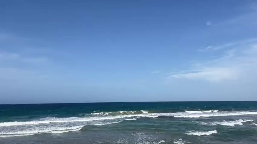 This is a wide shot of a beach and the ocean under a clear blue sky with some wispy clouds. Gentle waves are breaking on the shore, creating white foam. The water is a deep blue-green color. The sand of the beach is visible on the right side of the frame. The sun appears to be high in the sky, casting bright light on the scene. There are no people or objects visible in the frame.