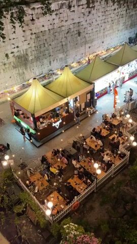 The video pans across a nighttime scene with several outdoor dining areas and market stalls set up along a riverbank. In the foreground, tables and chairs are occupied by diners, illuminated by string lights. Beyond this, market stalls with green awnings are visible, also lit and with people browsing. The river is dark, reflecting the lights from the opposite bank, where buildings and a bridge are visible. The scene suggests a lively evening atmosphere with food vendors and people enjoying themselves outdoors.