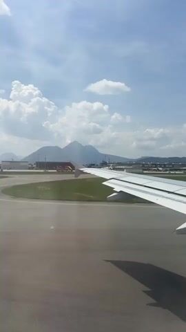 The video captures the view from an airplane window, likely during takeoff or landing at Salzburg Airport in Austria. The primary subjects are the airplane's wing, the airport tarmac, a grassy area, and a control tower in the distance. Mountains form a picturesque backdrop under a bright blue sky dotted with fluffy white clouds.

The activity appears to be airport operations, with the plane on the runway, suggesting an imminent departure or arrival. The time of day is clearly daytime, with bright sunlight casting shadows. The weather seems pleasant, with clear skies and sunshine.

Notable details include the prominent white airplane wing extending into the frame, the grey tarmac with yellow markings, and the lush green grass. The control tower, a tall structure with windows, is a distinct feature of the airport environment. The mountains in the background are impressive and add to the scenic beauty of the location. The overall impression is one of a typical airport scene in a visually appealing, mountainous region. No discernible text is visible in the video.